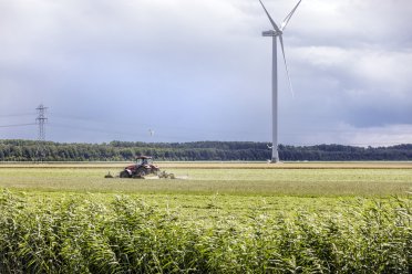 Afbeelding van een tractor die het gras maait in de buurt van een windmolen.