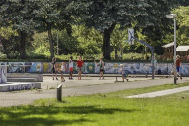 Afbeelding van een groepje kinderen die op een basketbalveldje met een bal spelen en kinderen met een skateboard en een step.